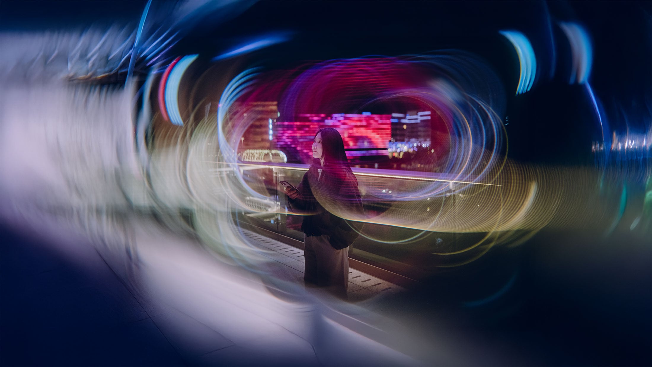 A woman standing on a bridge at night, illuminated by colorful, swirling lights and digital displays in the background, creating a dynamic, futuristic atmosphere.