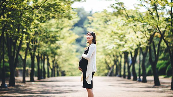 A pregnant woman stands on an outdoor pathway in the sunshine.