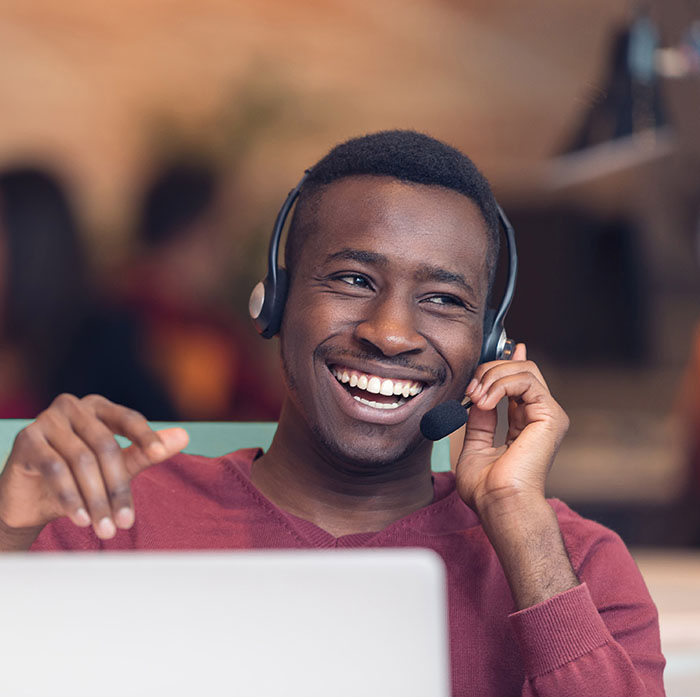 man with headset smiles. he is sat in front of a laptop.