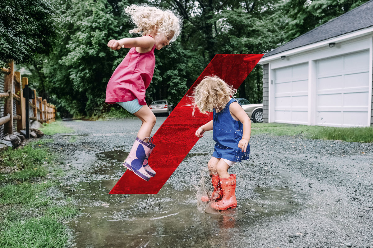 Two young children jumping and splashing in a puddle on a driveway in front of a house surrounded by green trees.